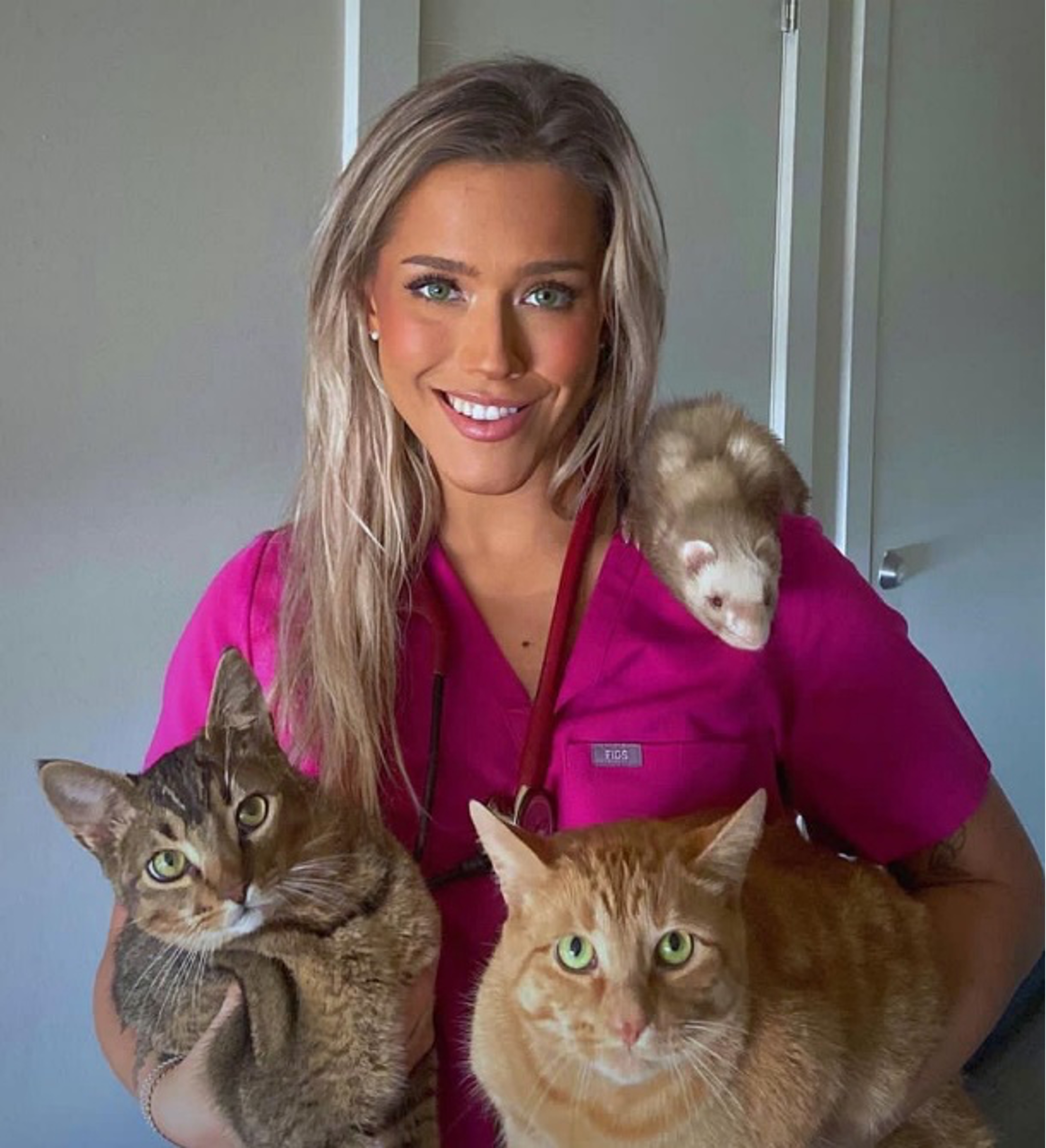 Ava is smiling at the camera. She is holding 2 large cats, one is grey and one is orange. There is a ferret on her shoulder.