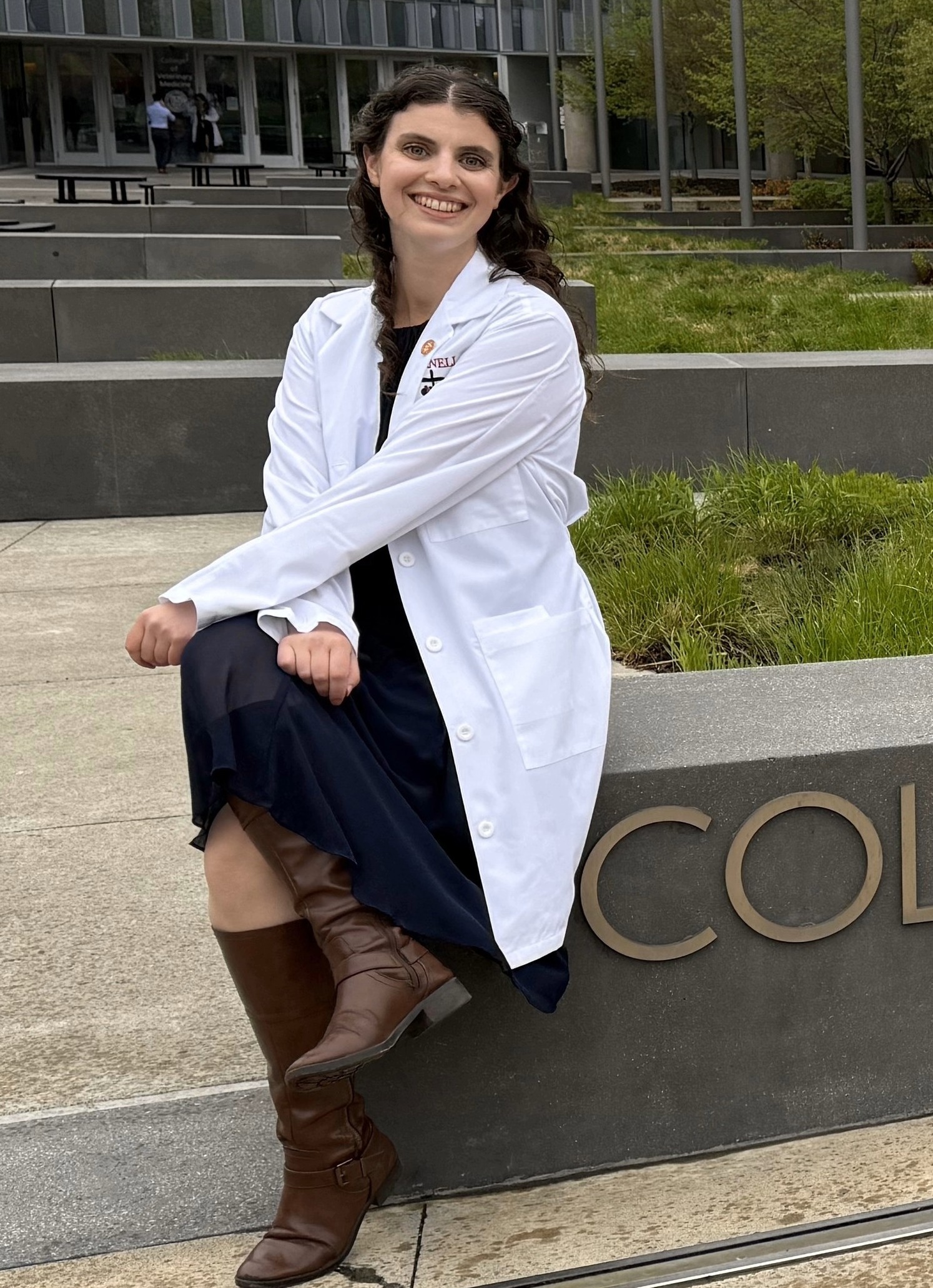 Danielle is sitting on a concrete bench wearing a white lab coat and smiling at the camera.
