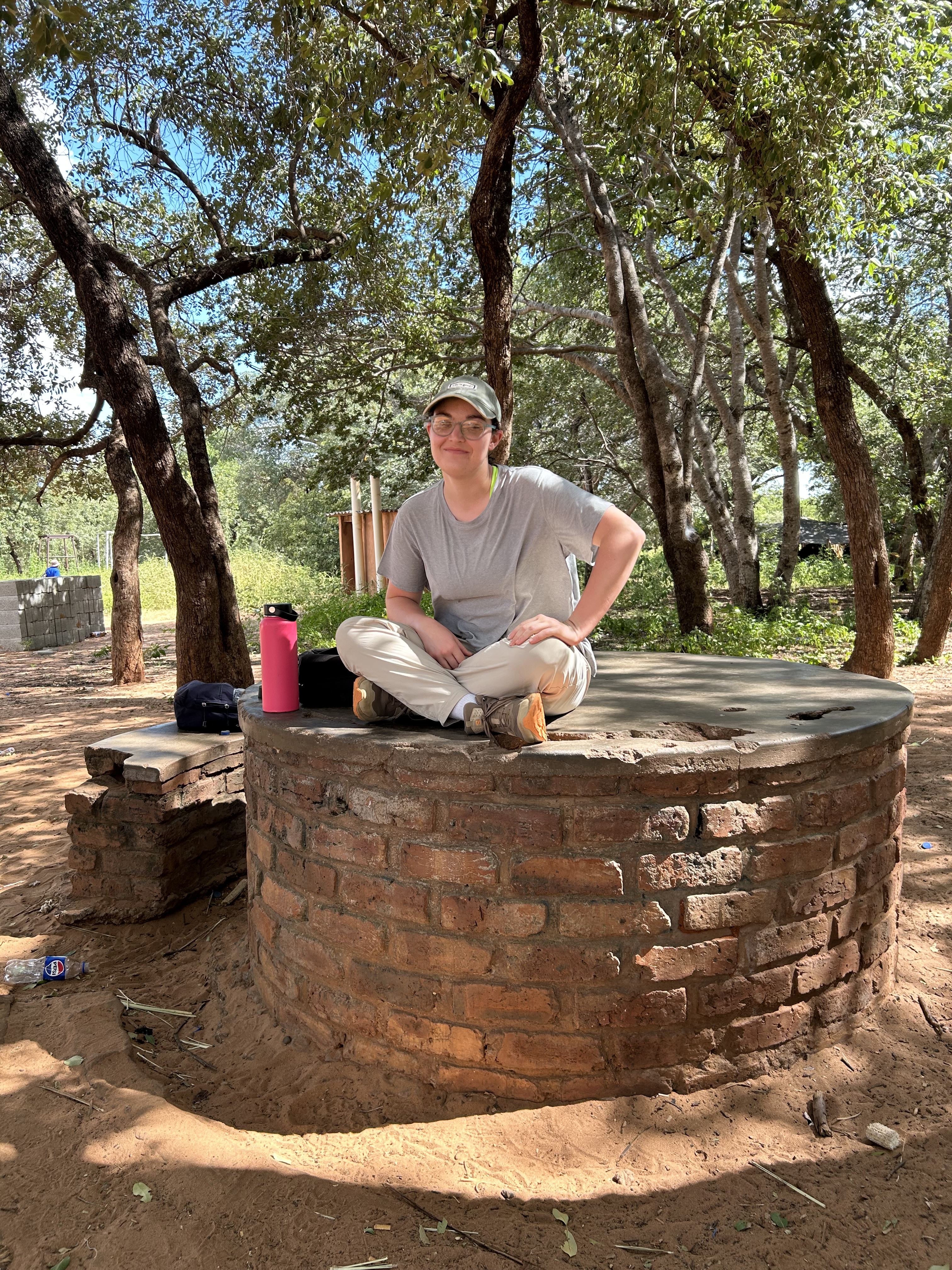 Brenna is sitting, cross-legged, on a brick structure and smiling at the camera
