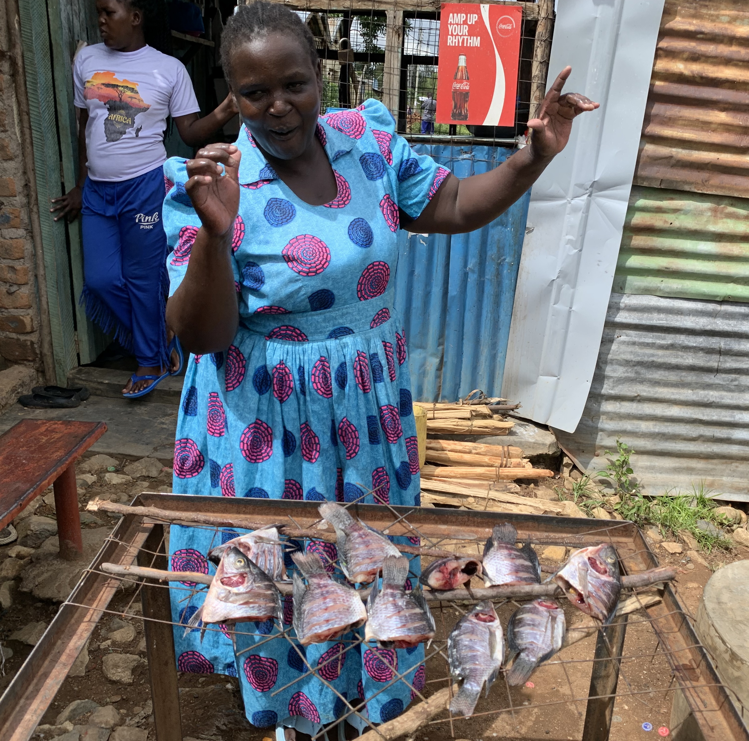 A smiling woman wearing a bright blue dress with pink circular patterns gestures while standing behind a grill displaying several freshly prepared fish. She is outdoors beside a corrugated metal structure, with another person and a “Coca-Cola” sign visible in the background.