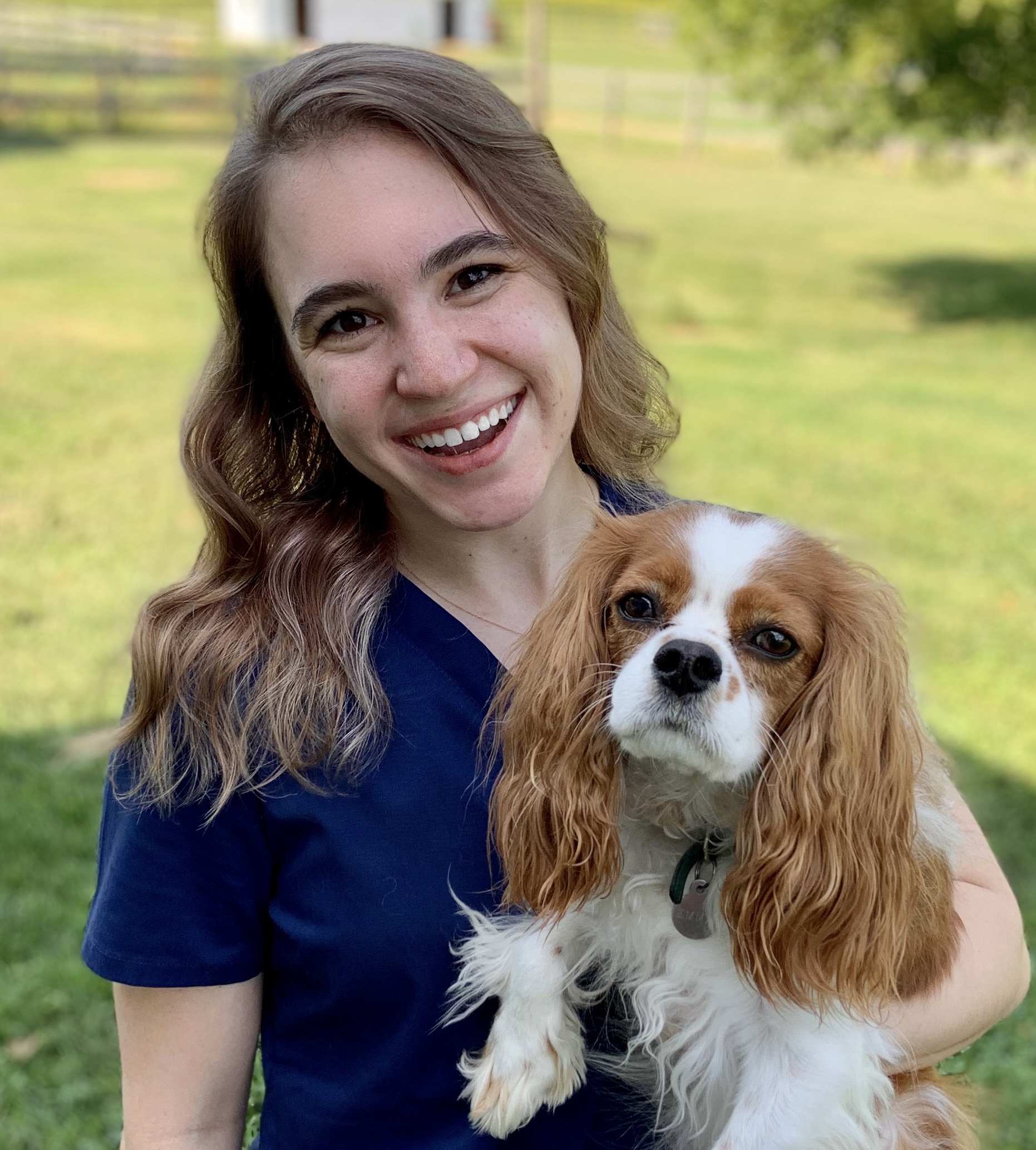 Shayna Orens is smiling at the camera with a King Charles Cavalier Spaniel sitting on her lap.