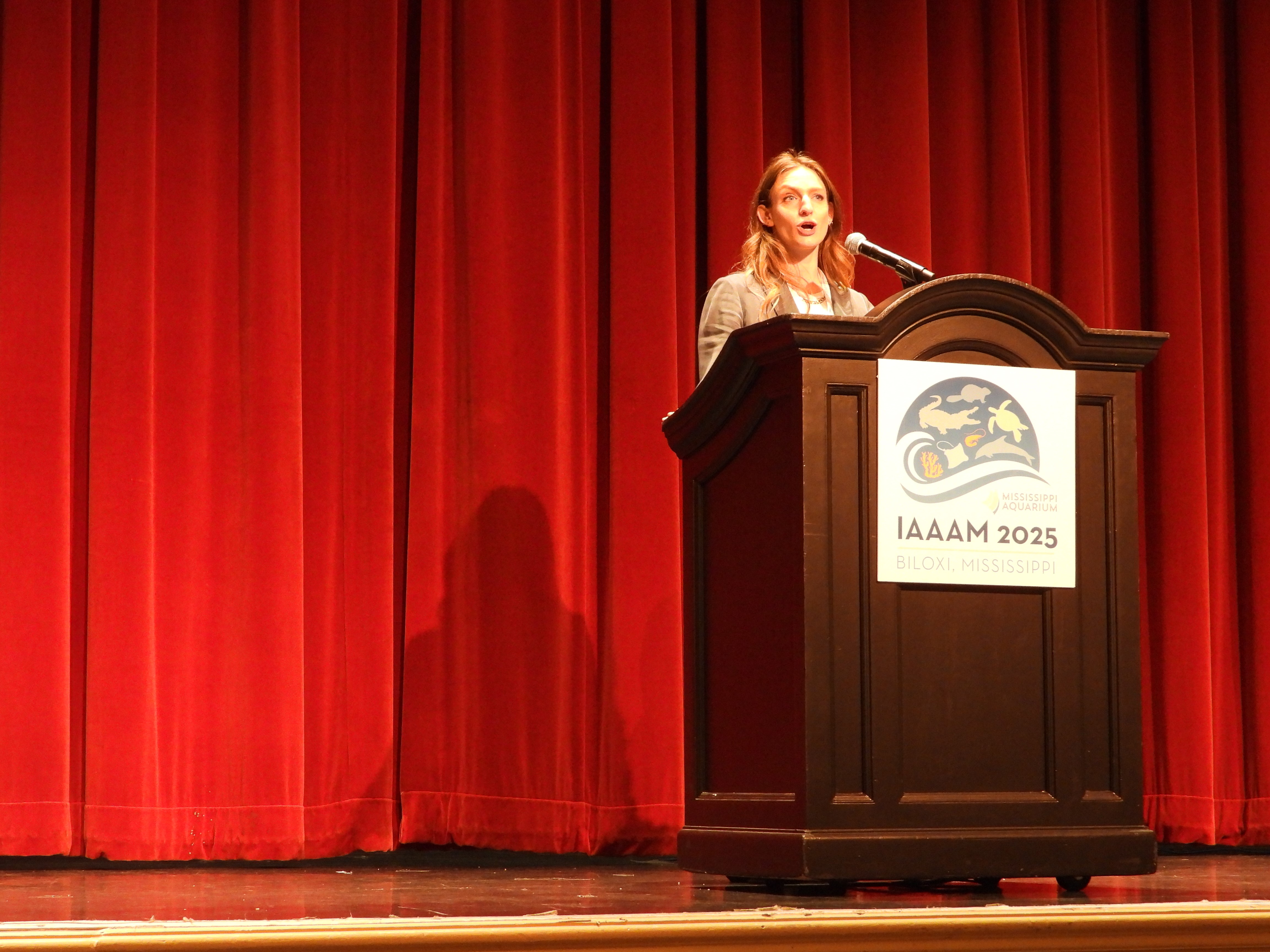 Emma is speaking at a podium with a red curtain in the background. The podium has a sign that says "IAAAM 2025".