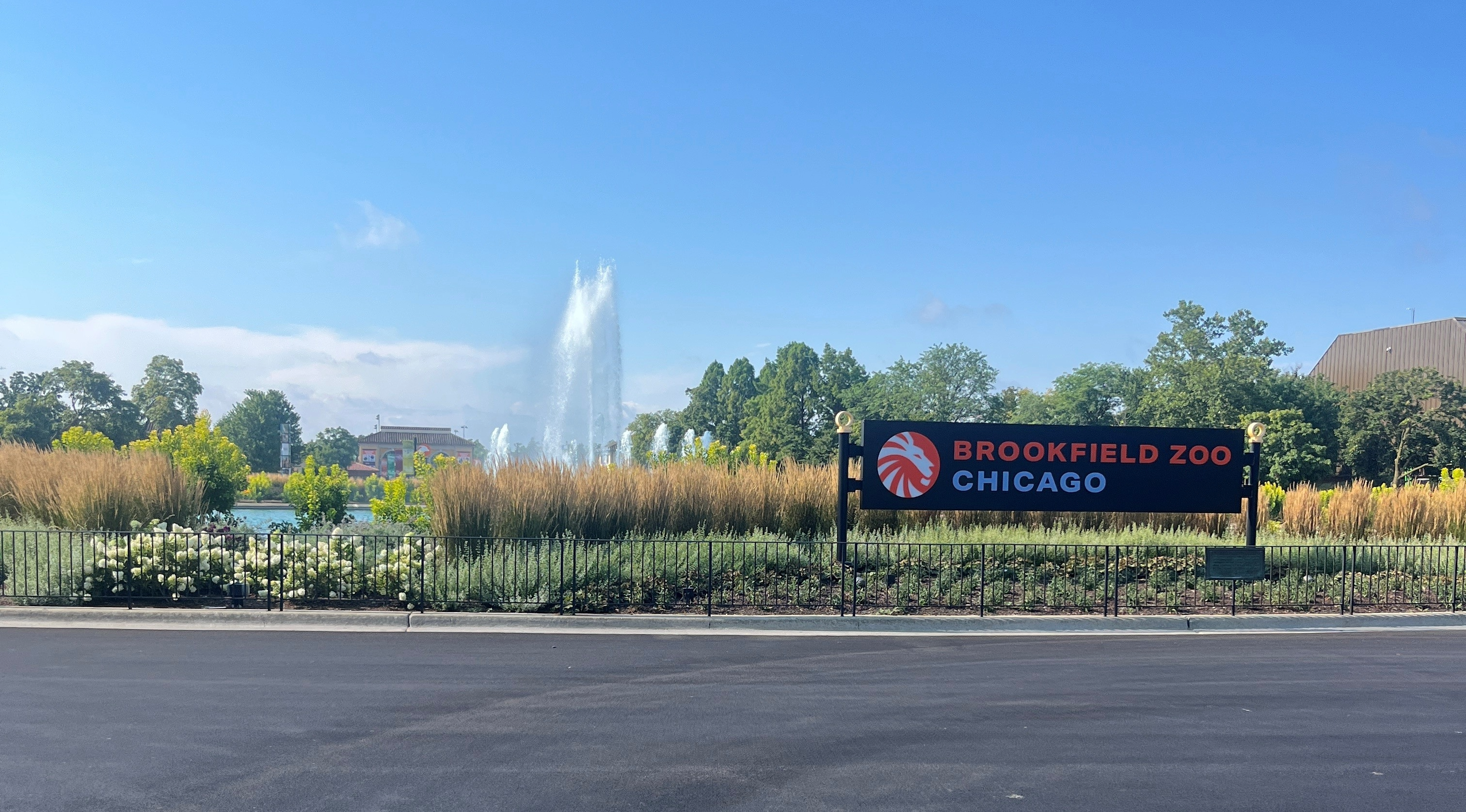 Brookfield Zoo Chicago entrance sign beside landscaped gardens, with a large fountain and trees in the background under a clear blue sky.