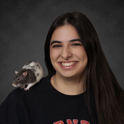 Ishita Monga smiling at the camera with a rat on her shoulder.