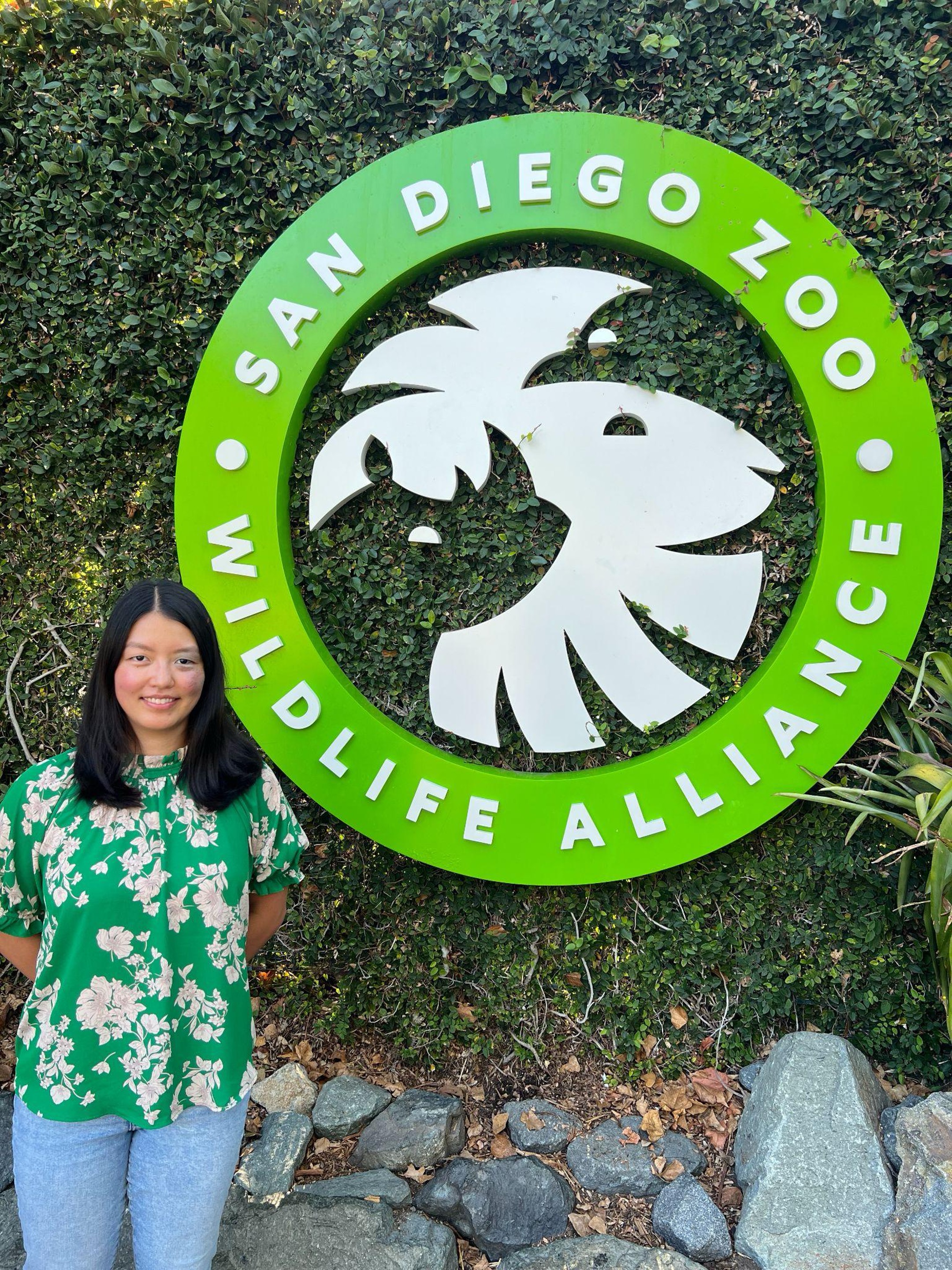 Kieran standing next to a logo featuring a rhino and lion that says "San Diego Zoo Wildlife Alliance".