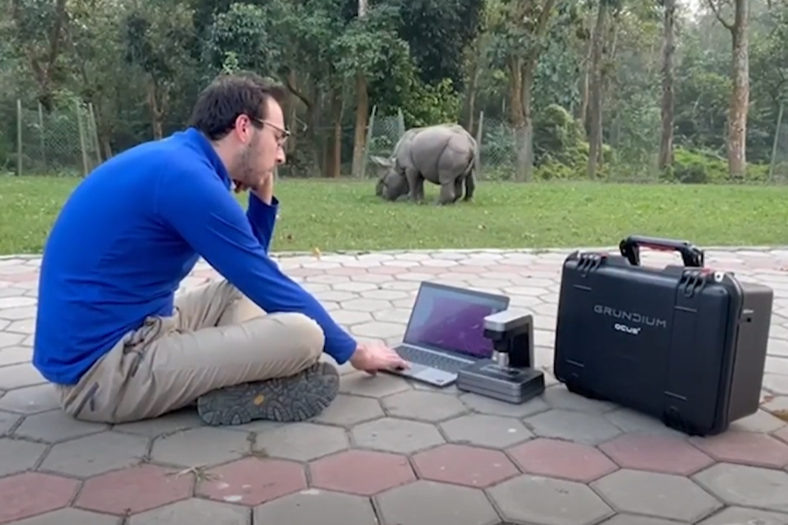 Carmen Smith using a laptop computer and a Grundium slide scanner to review slides while a juvenile rhino grazes in the background.