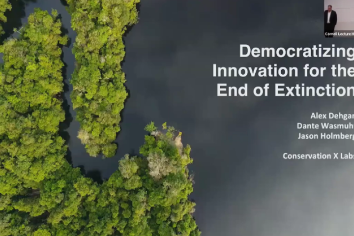 Aerial view of dense green forest and winding dark river, with overlaid presentation title text: “Democratizing Innovation for the End of Extinction” by Alex Dehgan, Dante Wasmuht, and Jason Holmberg of Conservation X Labs. A small inset in the upper right shows a lecture hall scene with a speaker at a podium, labeled "Cornell Lecture Hall."