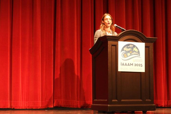 Emma is speaking at a podium with a red curtain in the background. The podium has a sign that says "IAAAM 2025".