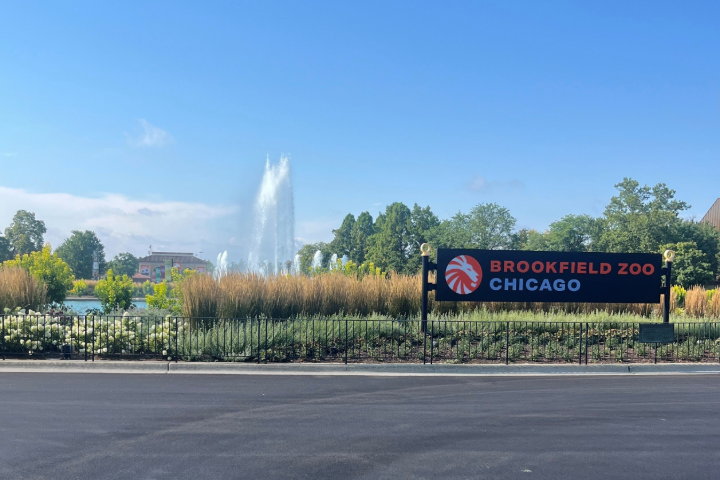 Brookfield Zoo Chicago entrance sign beside landscaped gardens, with a large fountain and trees in the background under a clear blue sky.
