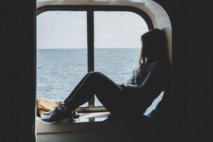 Person sitting by a large window, looking out at the ocean from inside a dimly lit cabin or ferry.