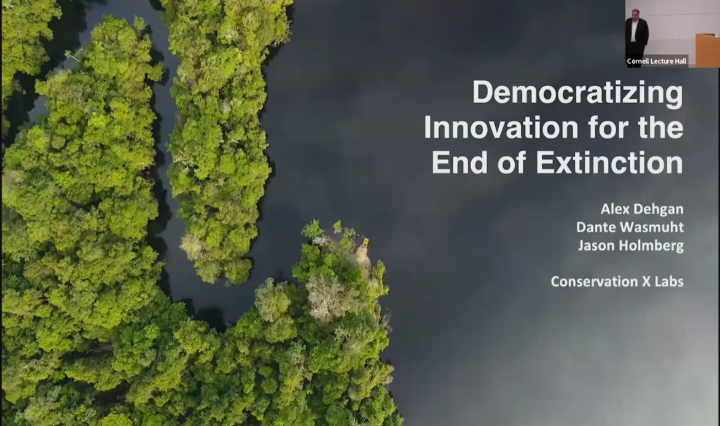 Aerial view of dense green forest and winding dark river, with overlaid presentation title text: “Democratizing Innovation for the End of Extinction” by Alex Dehgan, Dante Wasmuht, and Jason Holmberg of Conservation X Labs. A small inset in the upper right shows a lecture hall scene with a speaker at a podium, labeled "Cornell Lecture Hall."