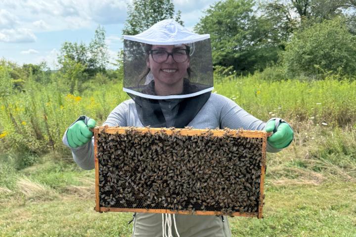 Taylor is wearing a protective suit and smiling while holding a beehive outside.
