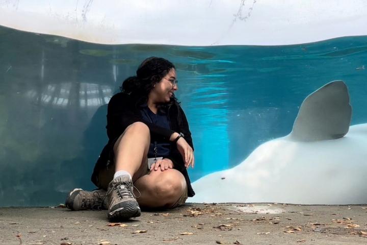 Natalia sits cross-legged on the ground and smiles toward a beluga whale resting against the glass of a large aquarium tank.