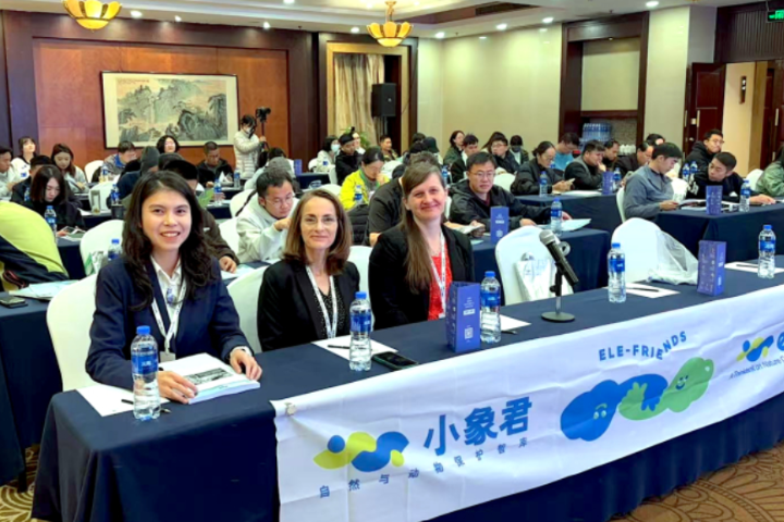 Three individuals sitting at a table with several people at tables behind them in a ballroom/conference setting.