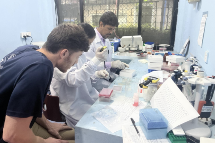 Zach and two other people wear white lab coats while sitting at a desk with various lab samples.